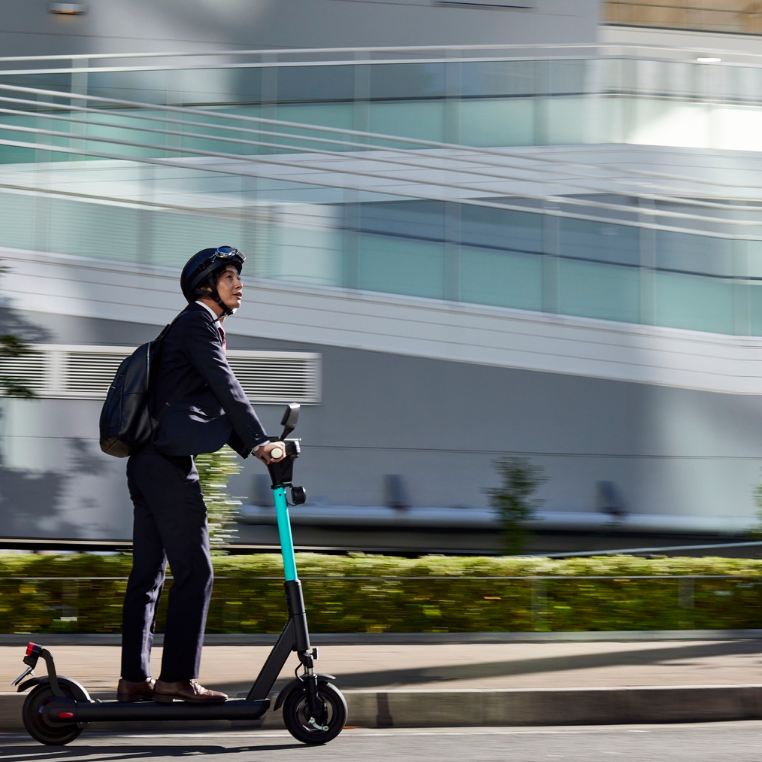 Mann in Anzug und Helm fährt auf einem E-Scooter entlang einer Straße.